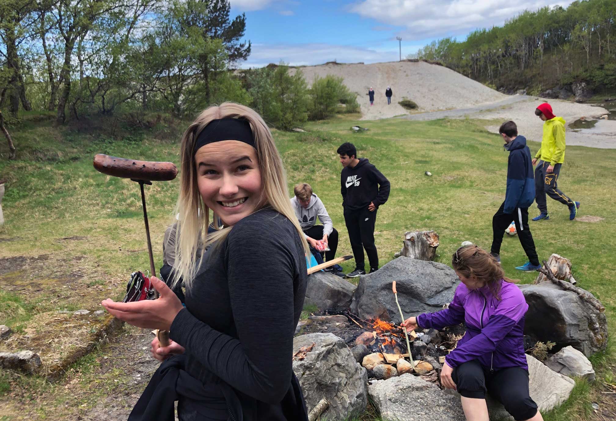 Bilde av en klasse på tur ute, fokuset er på en jente som har grillet seg en pølse på et spyd, hun smiler - Kristen Videregående Skole Nordland - E-sport