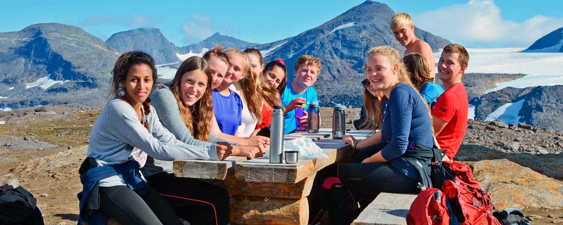 Bilde av en gjeng elever, gutter og jenter, på fjelltur med klassen, de sitter ved et trebord på benker i tre, det er turklær og utstyr rundt dem, termoser på bordet, de er i fjellene på Nesna - Kristen Videregående Skole Nordland - E-sport