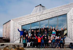 Bilde av elever, lærere og rektor på en klassetur på fjellet, det er foran en stor hytte med store vinduer, alle rekker hendene i været og smiler til kamera - Kristen Videregående Skole Nordland - E-sport