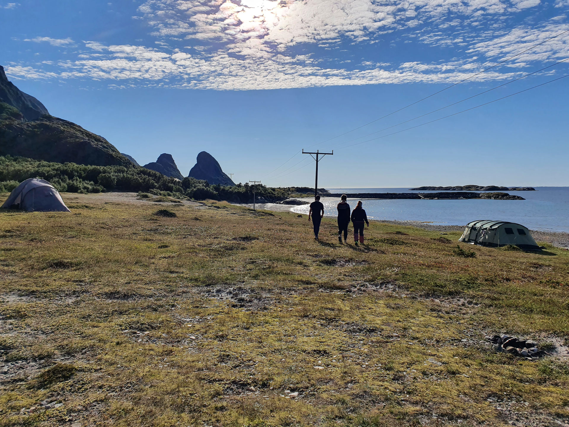 Bilde av stort fjordlandskap med vann, fjell og grønn bakke, det er satt opp to telt på hver sin ende av bildet, det er tre elever som går sammen vekk fra bildet og lengre inn i bildet - Kristen Videregående Skole Nordland - E-sport
