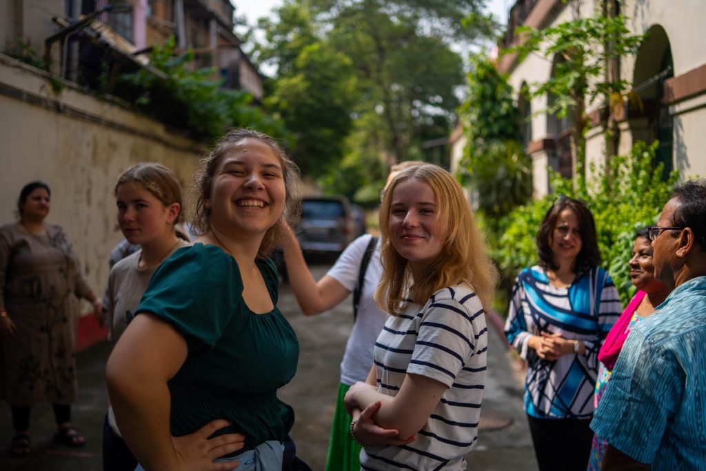 To elever som ser i kamera mens de står i en bakgård med masse grønne trær i bakgrunnen, noen andre mennesker står rundt