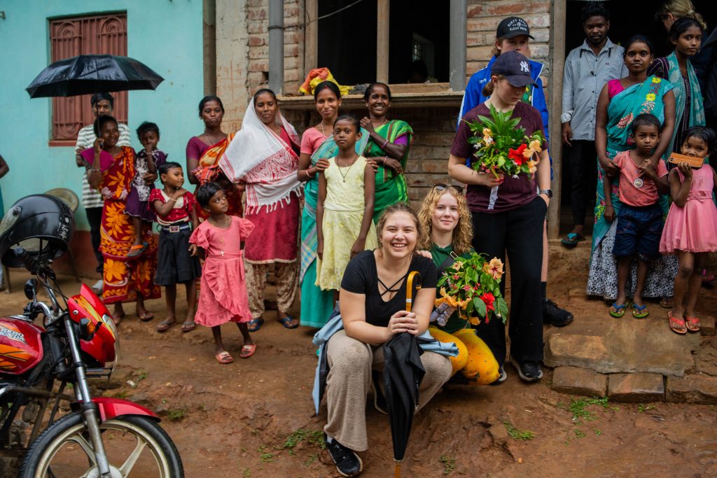 Bilde av to elever som sitter på huk sammen med innbyggere i en landsby i India