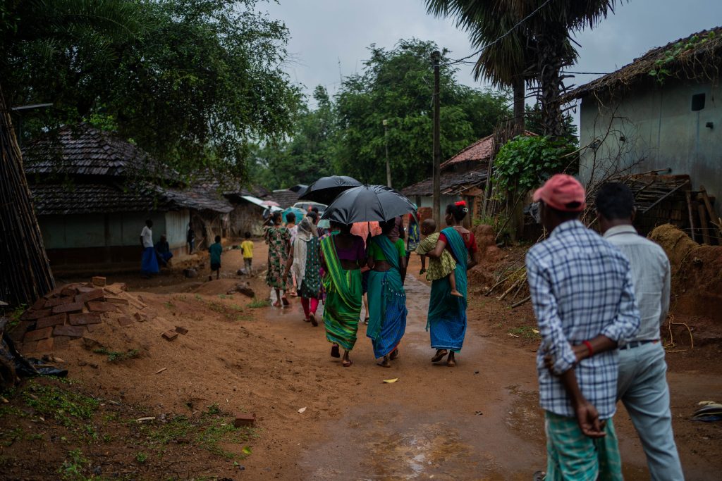 En gate i bygd i India. Tre damer i fargerike klær går bort fra kamera to av de under samme paraply