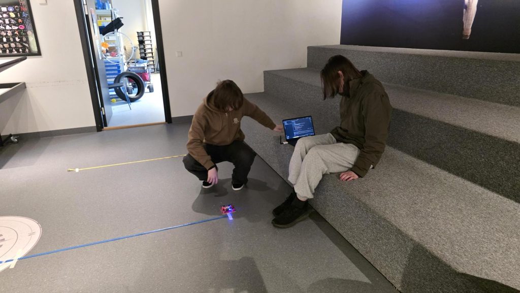 To elever som sitter rundt en drone som ligger på bakken, med en datamaskin like ved