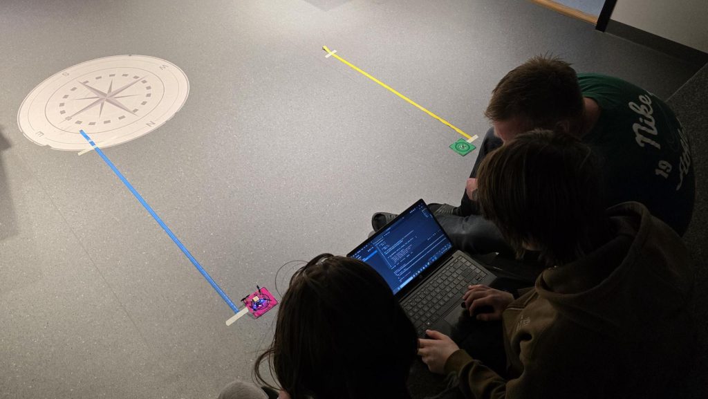 Tre elever som sitter rundt og ser på en pc. På gulvet ligger det to målebånd i to forskjellige farger