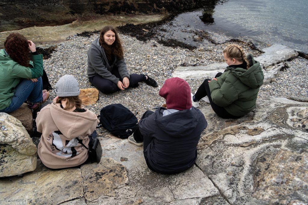 Elever som sitter samlet i fjæra på bergsteiner