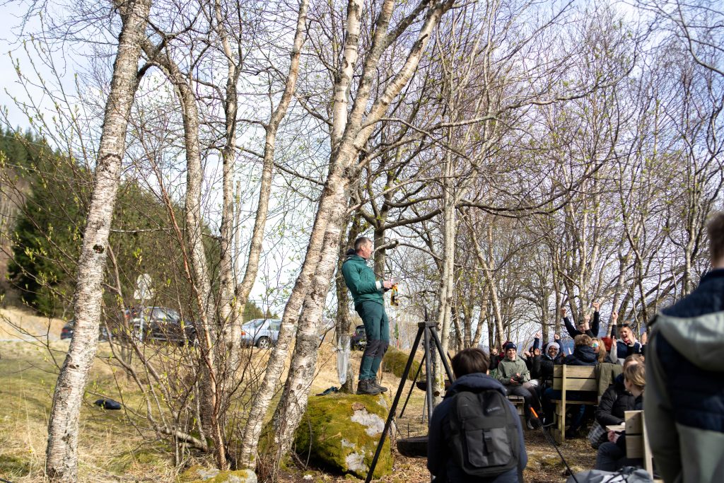 En voksen mann står på en stein omringet av bålbanne og bord fylt med elever som jubler i bakgrunnen