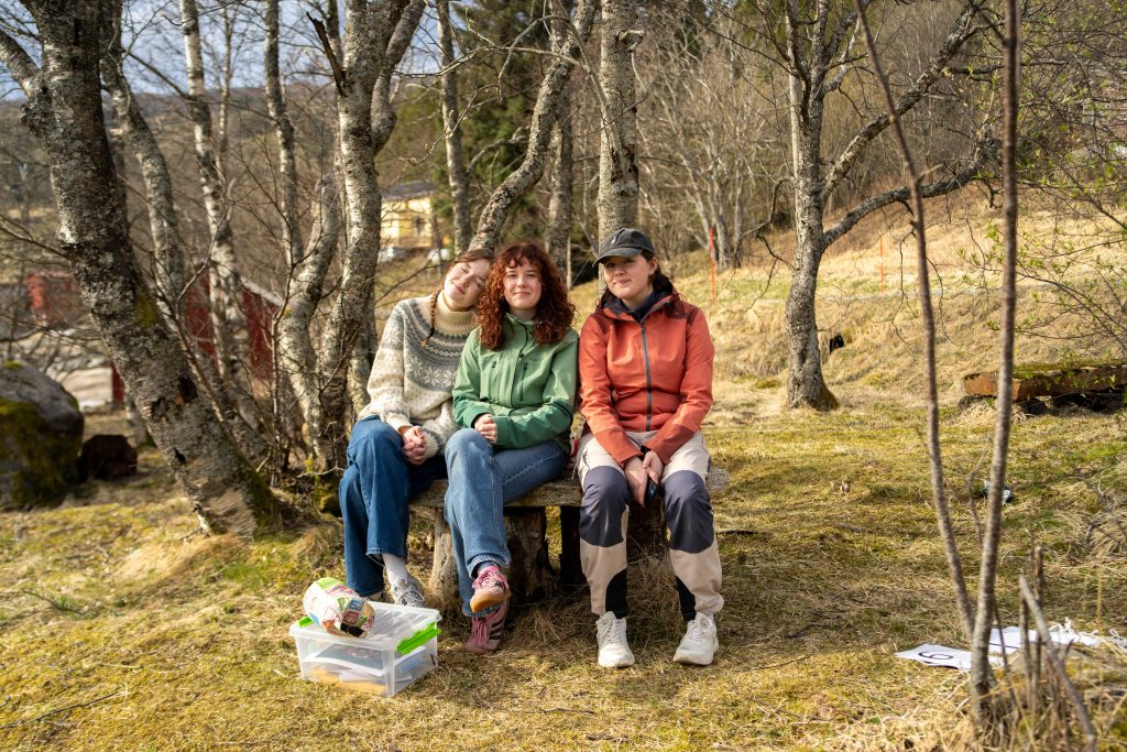 Tre elever som sitter sammen på en benk ute, og smiler til kamera